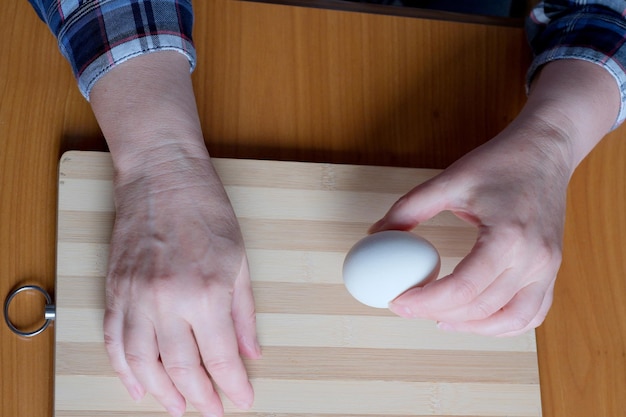 Mains d'une femme âgée montrant un œuf à la coque blanc et le pelant dans la cuisine à une table marron sans visage en gros plan