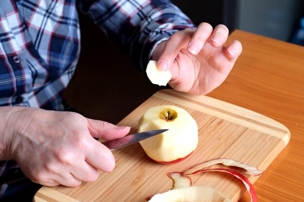 Photo les mains d'une femme âgée pèlent une pomme juteuse savoureuse et mûre rouge avec un couteau dans la cuisine à une table brune sans visage en gros plan