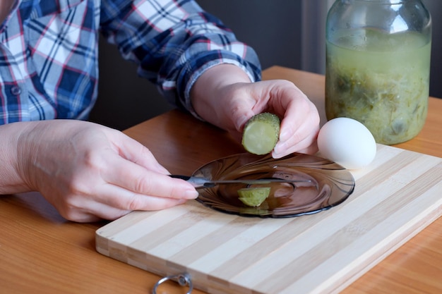 Les mains d'une femme âgée sortent de délicieux concombres marinés faits maison dans un pot pour les couper comme collation dans la cuisine sur une table brune sans gros plan du visage