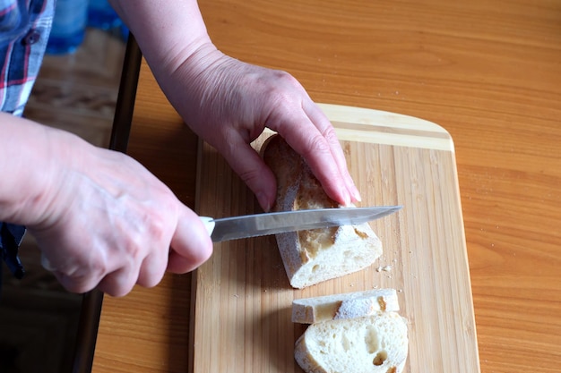 Mains d'une femme âgée tenant une baguette de pain de blé blanc en tranches dans la cuisine à une table marron sans visage en gros plan