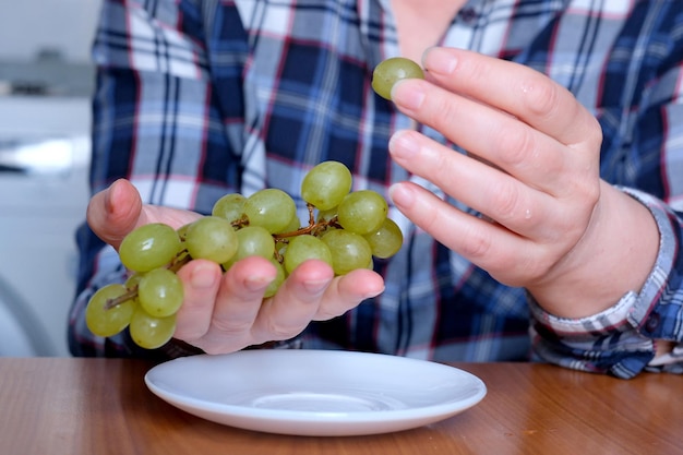 Les mains d'une femme âgée tiennent une petite brosse de raisins blancs sans pépins et les exposent dans la cuisine sur une table marron sans gros plan sur le visage