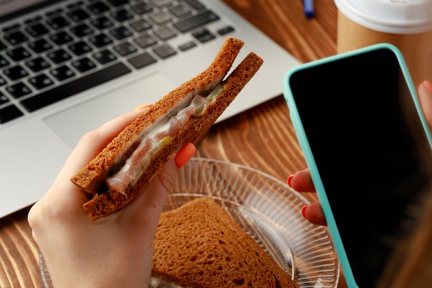 Mains d'une femme à l'aide de smartphone tout en ayant une pause déjeuner avec sandwich close up