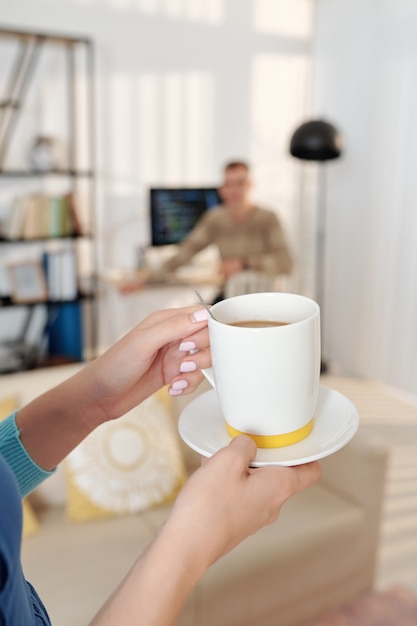 Mains de femme apportant une tasse de café à son mari qui travaille sur ordinateur à la maison en raison d'une pandémie