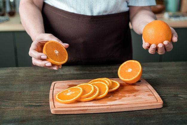 Mains de femme au foyer contemporaine en tablier debout par table de cuisine et tenant des oranges fraîches sur planche de bois avec des tranches juteuses