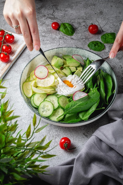 Les mains d'une femme coupent un œuf poché Salade verte avec des feuilles d'épinards de concombre d'avocat, des courgettes et des radis sur une table en pierre grise Menu du restaurant