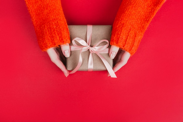 Photo les mains de la femme dans un pull tricoté chaud avec une boîte-cadeau de manucure enveloppée de papier kraft et d'un ruban rose sur le rouge.