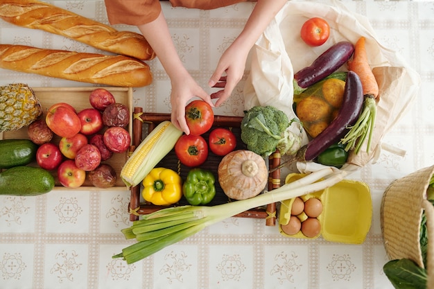 Mains de femme déballant des sacs recyclables de fruits et légumes à la table de la cuisine après son retour du marché, vue du haut