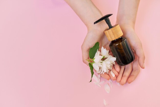 Mains de femme avec fleurs de jasmin et produit cosmétique en bouteille, cosmétiques bio naturels. Vue de dessus, fond rose, espace de copie