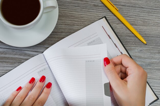 Mains de femme prenant une page de son bloc-notes. Tasse de thé, stylo sont en arrière-plan. Photo vue de dessus