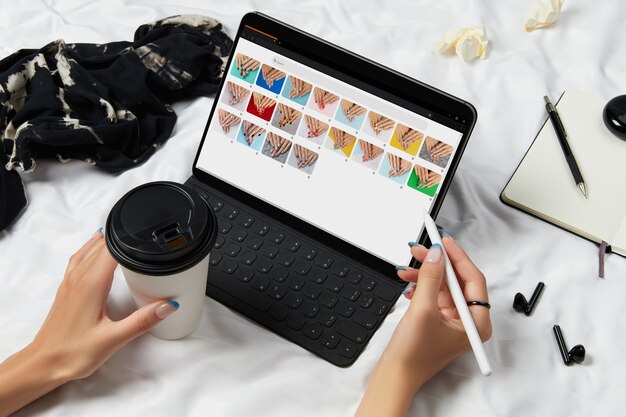 Photo mains de femme et tablette noire avec des dessins de manucure à l'écran sur un lit blanc