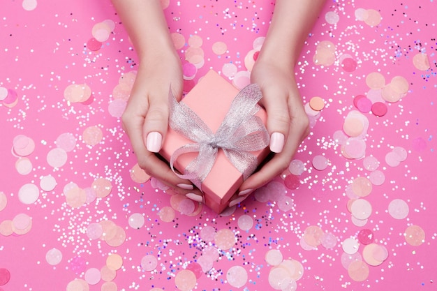 Mains de femme tenant une boîte cadeau avec un arc argenté sur fond rose pastel avec des paillettes et des confettis