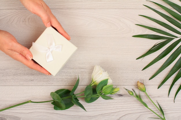 Mains de femme tenant une boîte-cadeau sur un fond en bois gris avec de belles fleurs et des feuilles vertes. Concept de donner un cadeau en vacances ou anniversaire. Vue de dessus.
