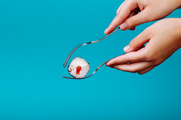 Mains de femme tenant un rouleau de sushi frais avec une fourchette, isolés sur fond bleu. Comment manger des sushis sans baguettes.