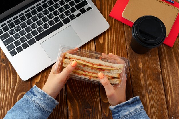 Mains d'une femme tenant un sandwich au-dessus de la table de travail avec ordinateur portable ouvert