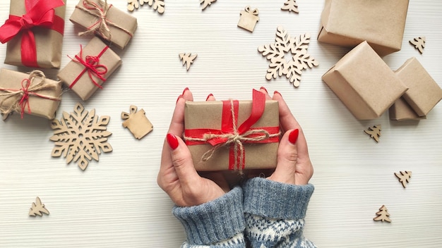 Les mains de la femme tiennent le cadeau de Noël dans du papier kraft avec un ruban rouge et une corde de jute. Table en bois avec des cadeaux de Noël et des flocons de neige en bois. Vue de dessus.