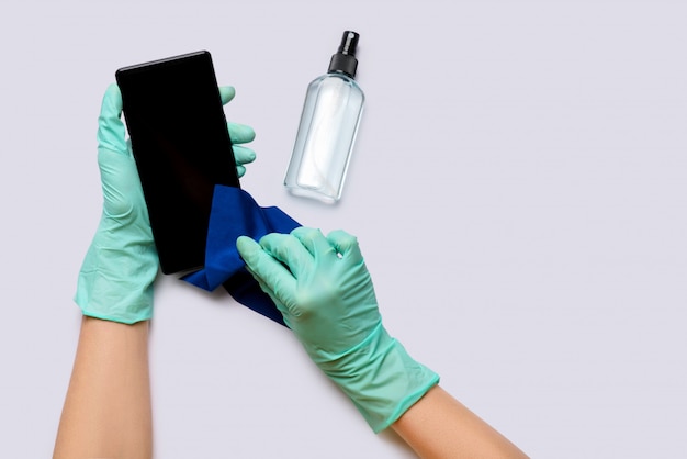 Les mains des femmes dans des gants en latex essuyant le téléphone mobile avec de l'alcool décontaminant la vue de dessus sur fond gris clair