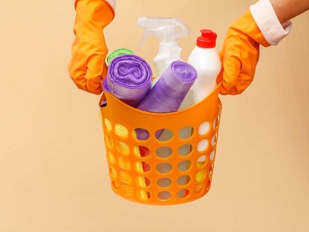 Les mains des femmes dans des gants de protection en caoutchouc tenant un panier orange avec des sacs à ordures, des bouteilles de nettoyant pour verre et carrelage, une éponge sur fond beige. Concept de lavage et de nettoyage.