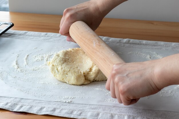 Les mains des femmes déroulent la pâte crue caillée avec un rouleau à pâtisserie en bois sur du papier sulfurisé sur une nappe en lin