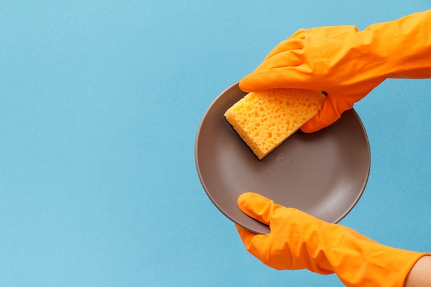 Les mains des femmes en gant de protection orange avec plaque et éponge sur fond bleu. Concept de lavage et de nettoyage.