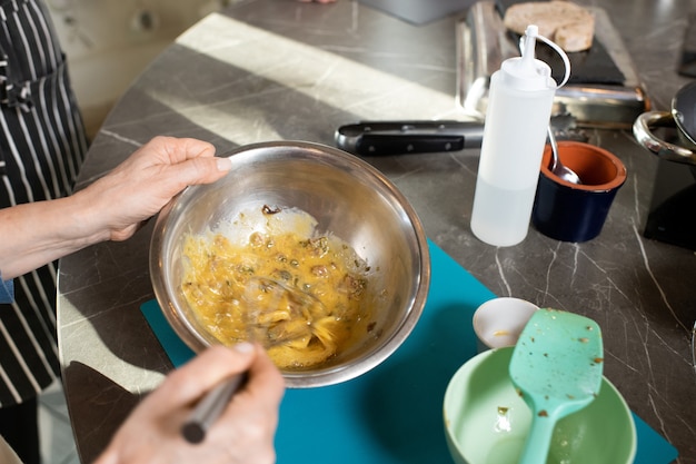 Mains de femmes matures mélangeant des ingrédients crus pour un ragoût avec des moustaches dans un bol métallique pendant la cuisson par table de cuisine à la master class