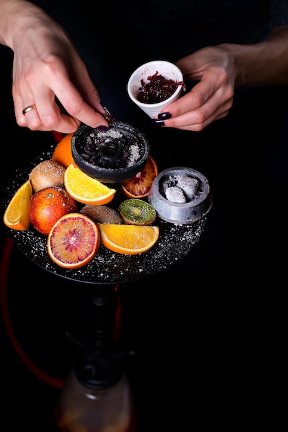 Les mains des femmes mettent du tabac aux fruits dans un narguilé sur un fond noir