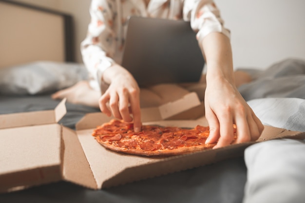 Les mains des femmes prennent un morceau de pizza dans une boîte, assis sur un lit à l'aide d'un ordinateur portable.