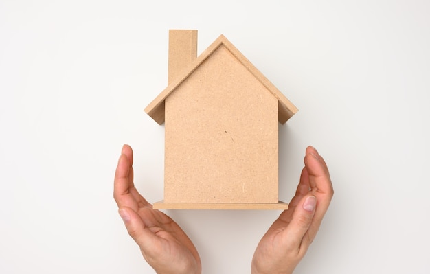 Les mains des femmes se sont jointes sur une maison modèle miniature en bois sur fond blanc. Concept d'assurance immobilière, protection de l'environnement, bonheur familial