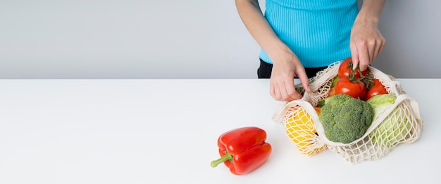 Les mains des femmes sortent des légumes frais d'un sac sur une table blanche Bannière