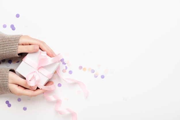 Les mains des femmes tiennent une boîte-cadeau sur une vue de dessus de fond blanc. La fille donne une surprise dans une boîte avec un ruban et des confettis. Concept de vacances, Noël, anniversaire, journée de la femme. Espace de copie.