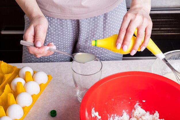 Les mains des femmes tiennent une cuillère et des bouteilles de jus de citron prêt à l'emploi et la versent dans la pâte à pétrir du fromage cottage et de la farine dans un bol rouge sur fond d'œufs dans un substrat jaune