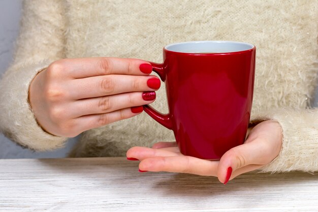 Les mains des femmes tiennent une tasse rouge dans le contexte d'un pull blanc moelleux avec manucure rouge