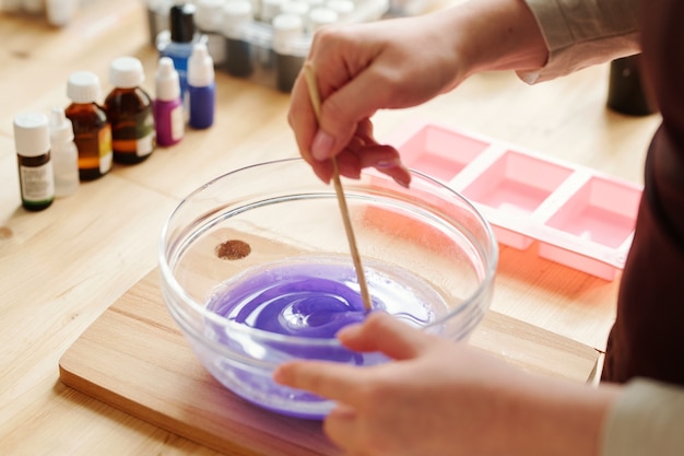 Photo mains de fille créative mélangeant la couleur lavande avec une masse de savon liquide dans de la verrerie avant de la verser dans des moules en silicone