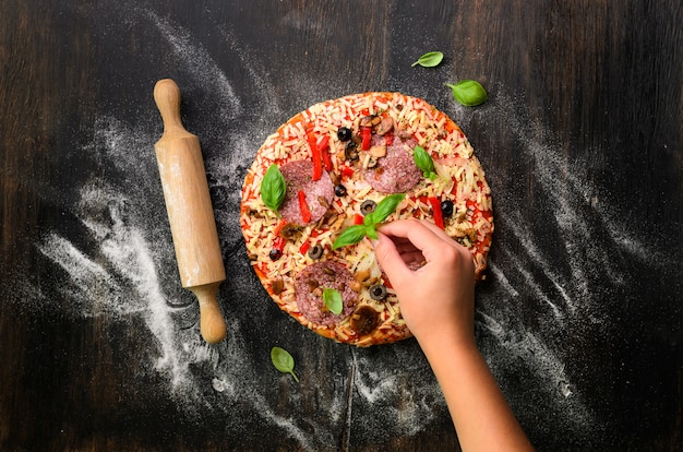 Mains De Fille Faisant, Décaper, Préparer Une Pizza Au Basilic Feuilles Sur Noir. Vue De Dessus, Espace De Copie