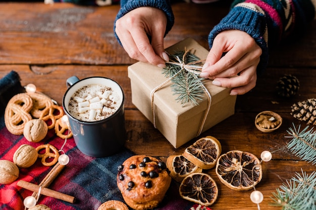 Mains De Fille Faisant Le Noeud Sur Le Dessus De La Boîte Cadeau Enveloppée Entourée D'aliments Sucrés, Boisson Chaude Dans La Tasse Et Les Noix