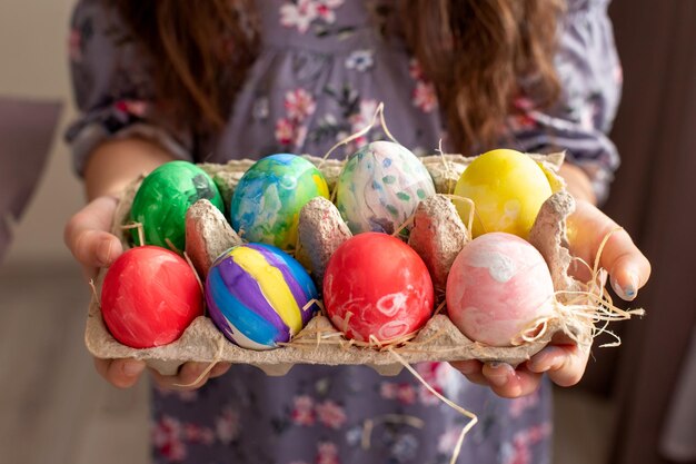 Mains d'une fille vêtue d'une robe tenant un plateau en carton avec des œufs colorés peints à la main