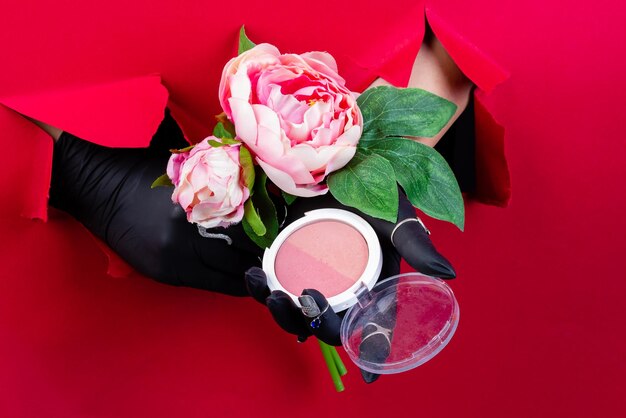 Mains gantées avec boîte à poudre ronde et fleurs traversant un mur de papier rouge