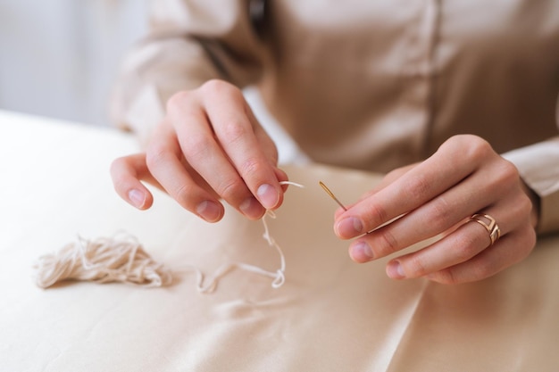 Mains en gros plan d'une femme méconnaissable mettant du fil dans le chas de l'aiguille assise à table. Gros plan d'une artisane mettant la mèche dans l'aiguille. Processus de fabrication de bougies naturelles faites à la main