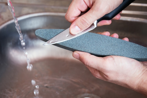 Les mains en gros plan d'un homme aiguisent soigneusement un couteau sous un jet d'eau sur une meule. Affûtage domestique à domicile. Entretien des couverts de cuisine.