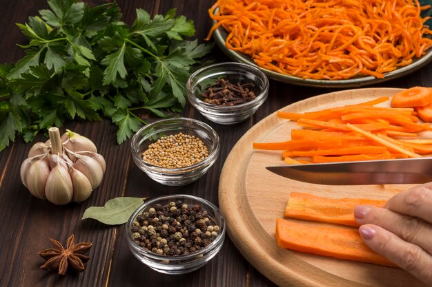 Les mains hachent les carottes crues sur une planche à découper. Épices, ail et persil sur table. Carottes fermentées en assiette. Source naturelle de renforcement de l'immunité. Fermer. Surface en bois foncé