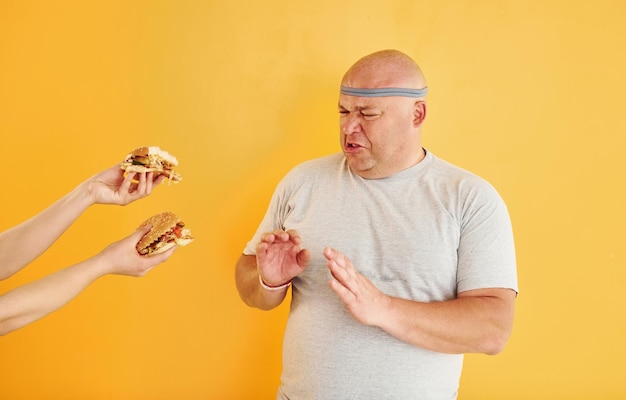 Les mains avec des hamburgers tentent le gars Un homme en surpoids drôle en cravate sportive est sur fond jaune