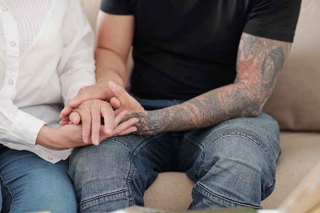 Mains d'homme aux bras tatoués tenant la main de sa mère mûre pour la rassurer et la soutenir