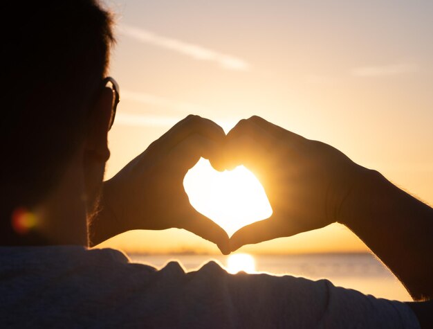 Des mains d'homme formant une forme de cœur avec une silhouette de coucher de soleil