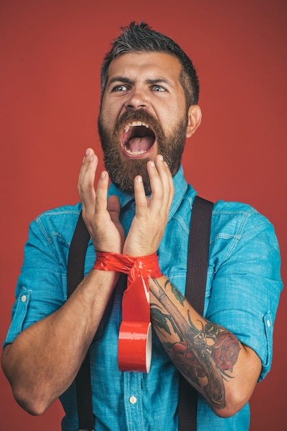 Mains d'homme liées avec du ruban adhésif rouge. Homme d'affaires barbu avec du ruban isolant sur les mains. Concept de danger, de peur, de choc et sans espoir - homme criant désespéré avec les mains attachées avec du ruban adhésif rouge. Isolé