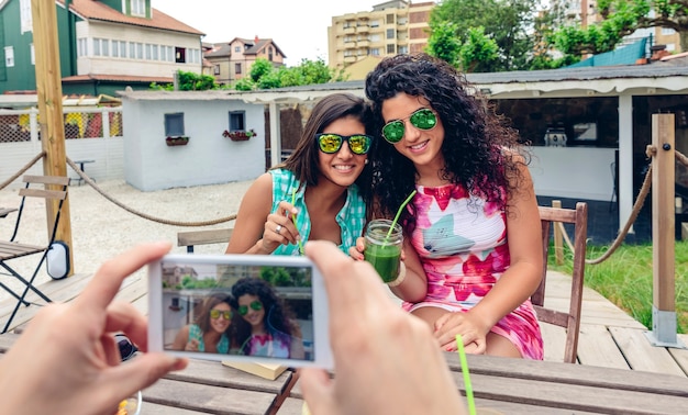 Photo mains d'homme prenant une photo avec un smartphone à deux jeunes femmes heureuses avec des lunettes de soleil tenant des smoothies aux légumes verts à l'extérieur