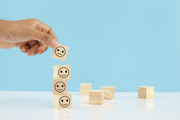 Les mains de l'homme ramassent des blocs de cube en bois de sélection avec des icônes d'émotion malheureuse sur fond bleu. personnel non sélectionné de l'entreprise. concept d'entreprise de notation et de classement.