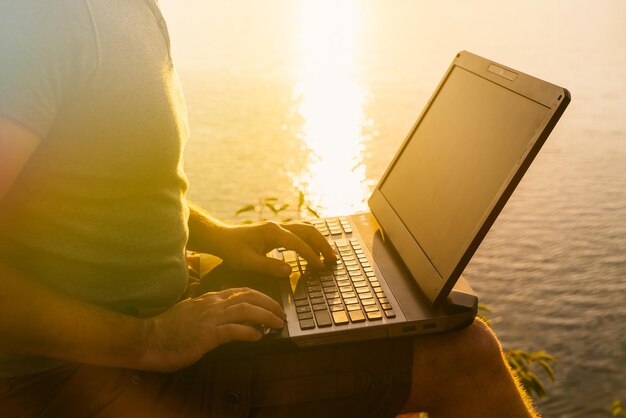 Les mains de l'homme tapent un message sur l'ordinateur portable près de la rivière au coucher du soleil Les rayons du soleil du soir tombent sur l'ordinateur portable de l'homme