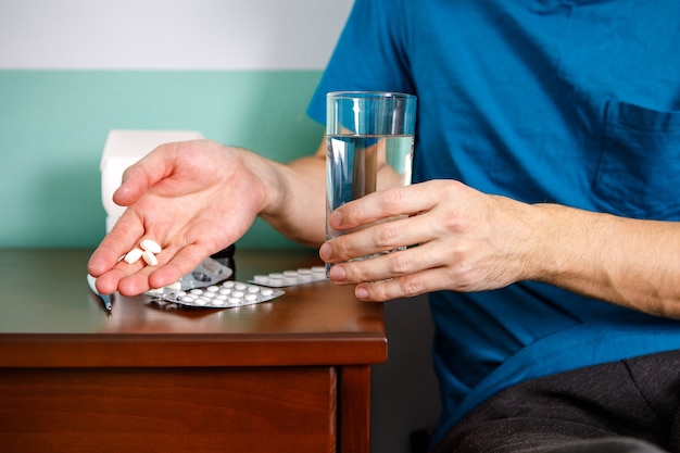 Mains homme en tenant la pilule et le verre d'eau sur la table alors qu'il était assis sur le canapé et va prendre des médicaments