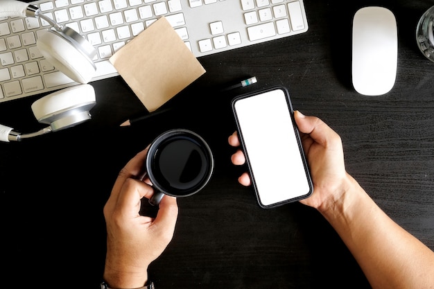 Mains d&#39;homme tenant une tasse à café et un téléphone portable sur un bureau sombre.