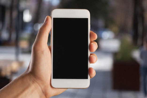 Les mains de l'homme tenant un téléphone portable avec un écran blanc sur le fond flou de la rue