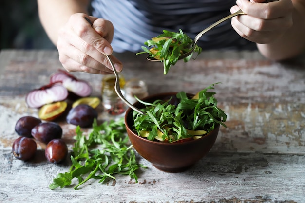 Les mains des hommes mélangent la salade dans un bol avec une cuillère et une fourchette. Salade légère à la roquette et aux prunes. Salade de prunes d'automne. Nourriture saine. Mise au point sélective.